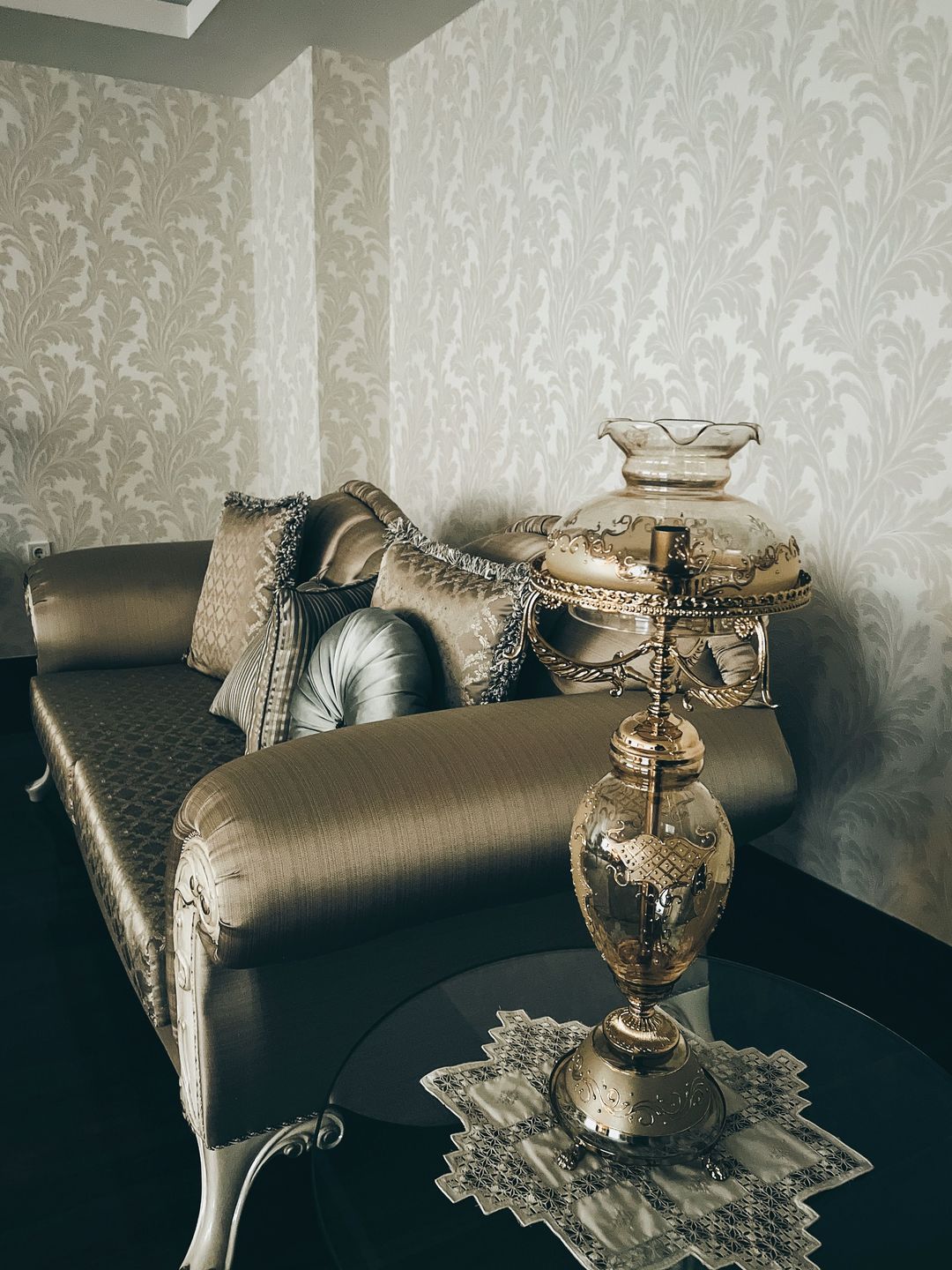 Moody sitting room with a pale scrolling damask wallpaper, a satin chaise and an ornate glass oil lamp