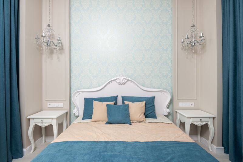 Symmetrical bedroom with a pale aqua damask wallpaper panel behind a carved white headboard, crystal sconces and teal curtains