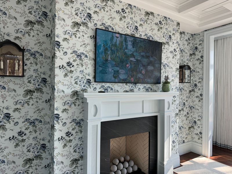 Living room papered in a blue-and-white floral wallpaper around a white fireplace mantel, with a framed water-lily painting above