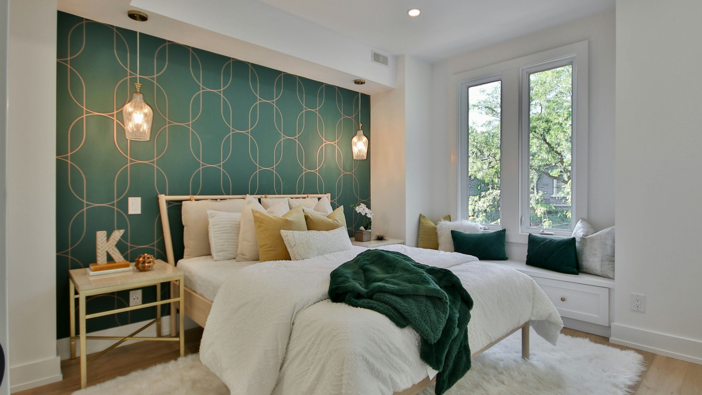 Bright bedroom with a deep green and gold geometric wallpaper accent wall behind the bed, glass pendant lights and a window seat