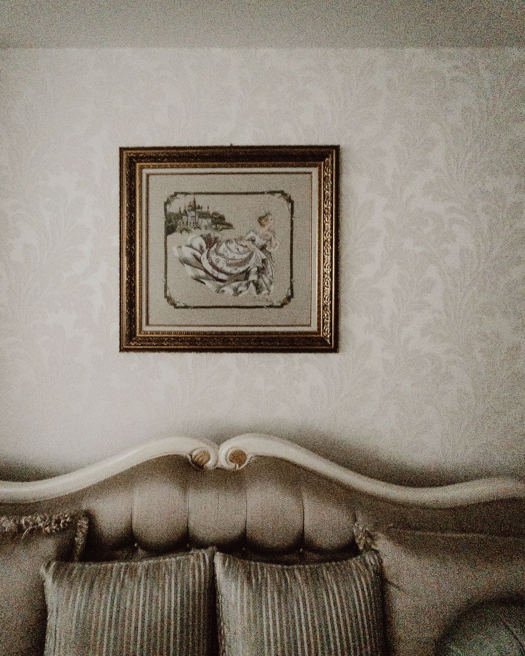 Close-up of ivory tone-on-tone damask wallpaper behind an upholstered headboard, with a small framed botanical print