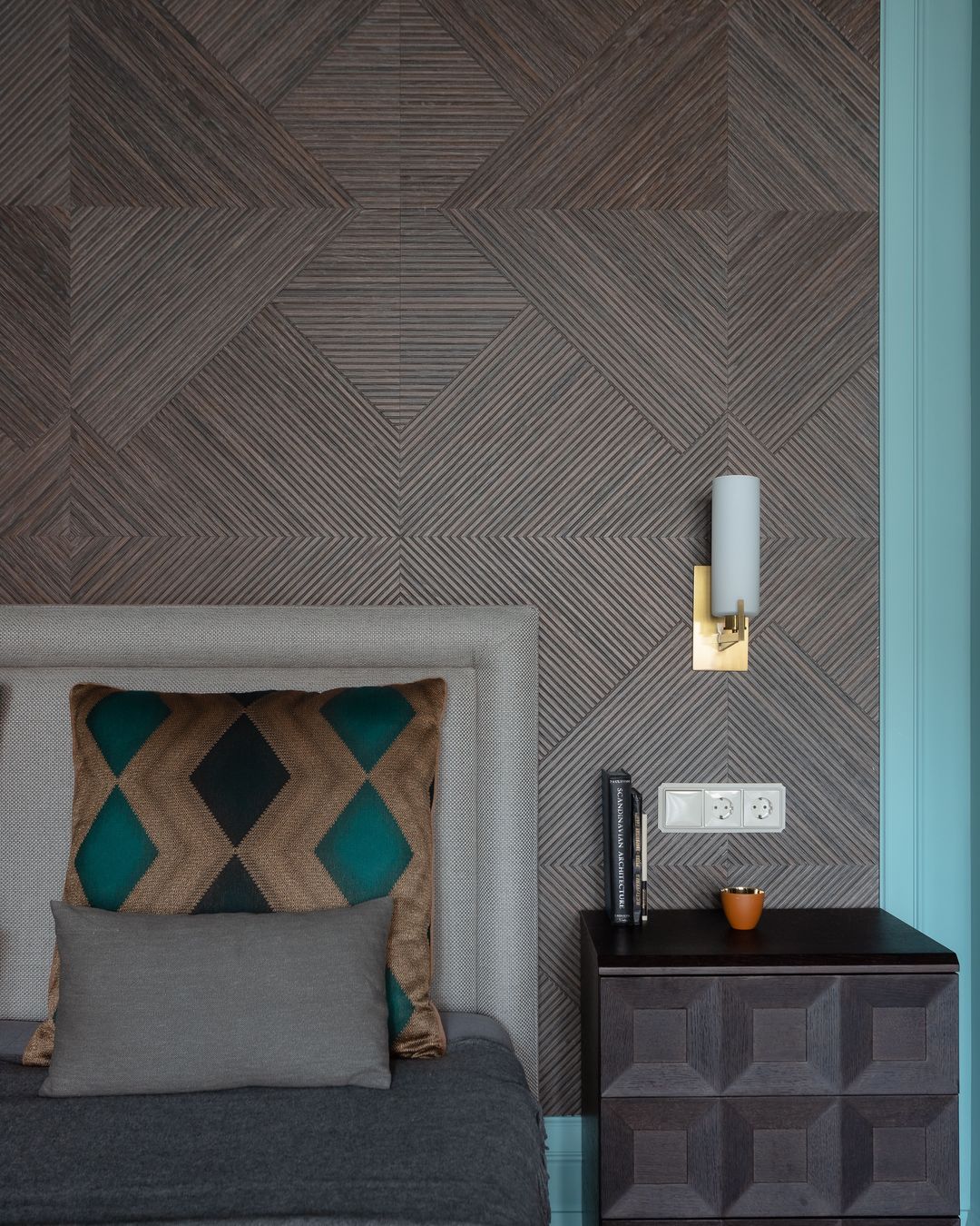 Bedroom wall covered in a dark geometric herringbone-style wallcovering with a brass wall sconce beside the bed