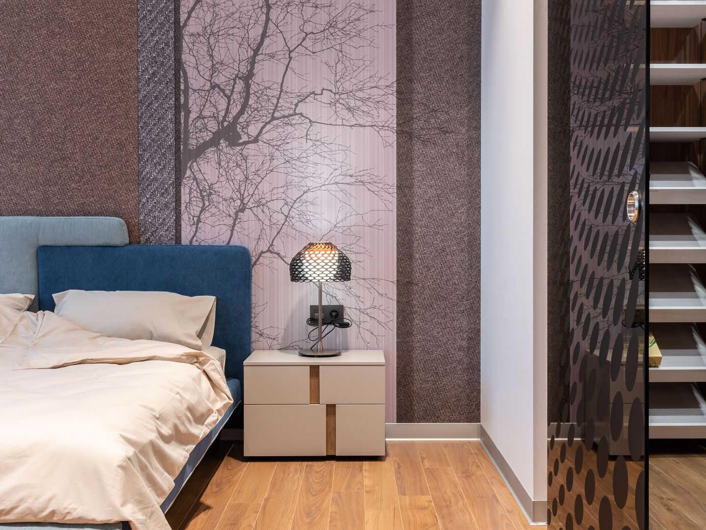 Modern bedroom with a soft lilac bare-branch mural panel between textured wallcovering strips, beside a blue upholstered bed